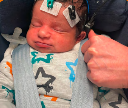 Newborn baby lying in a car seat or carrier, with small adhesive sensor pads placed on the head and a soft earpiece probe held just behind the ear by an adult hand, demonstrating a hearing test that measures the inner ear’s response to sound.