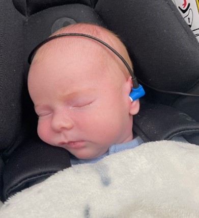 Babi yn cysgu mewn sedd car, gyda dyfais sain fach wedi’i gosod y tu ôl i’r glust a phlwg clust meddal yn y glust, wedi’i gysylltu â gwifren, gan ddangos sut mae prawf clyw baban newydd-anedig yn mesur sut mae’r glust fewnol yn ymateb i sain gan ddefnyddio offer arbenigol.
