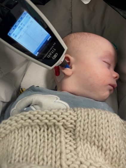 Sleeping infant with a medical monitor device near the head, screen visible beside them, wrapped in a chunky knit blanket in a care setting