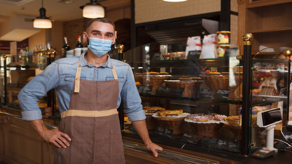 Bakery worker in a blue shirt and brown apron stands behind a glass pastry display, wearing a blue mask.
