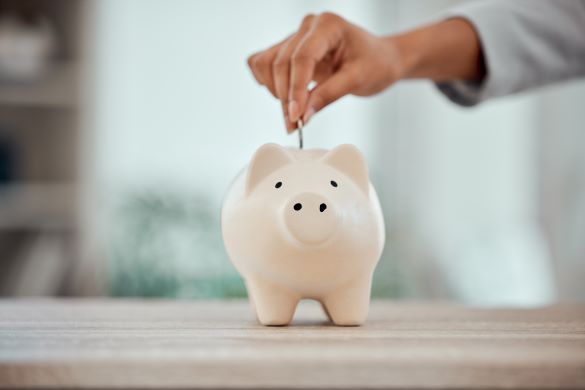 Hand placing a coin into a white piggy bank on a wooden table, symbolizing saving money.