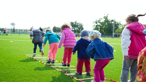 Children in colorful jackets stepping along a ground ladder on a sunny grassy field outdoors (outdoor activity/street-club style).