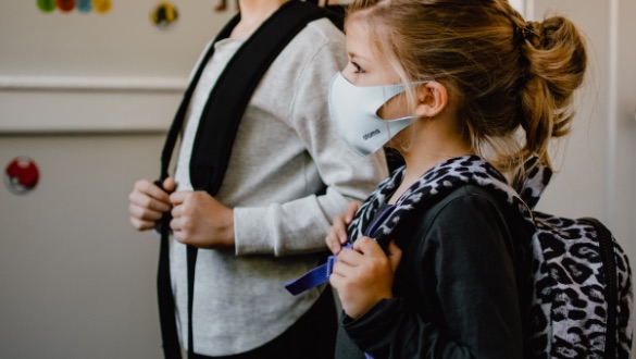 Two children wearing masks walk through a school hallway, each carrying a backpack. The girl in the foreground has a leopard-print backpack and a blue‑trimmed mask.