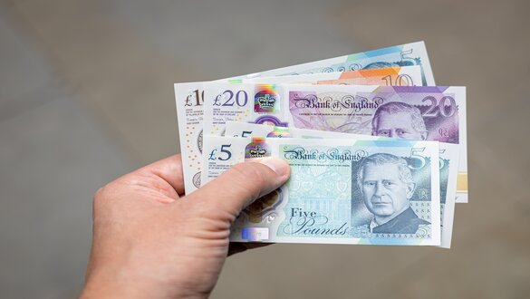 Hand holding a fan of Bank of England notes: £5 blue, £10 brown, £20 purple, and other denominations visible.