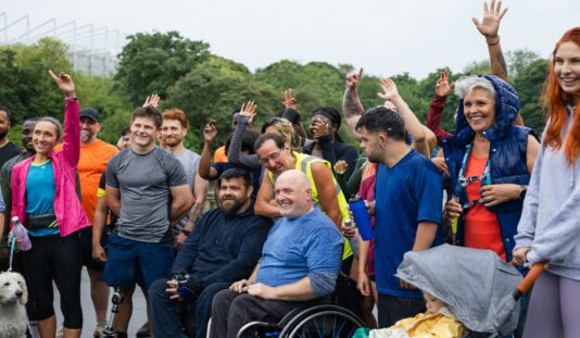 Diverse group of people outdoors at an event, many raising hands in the air; a man in a wheelchair sits near the center, a small white dog is on the left, and trees and a fence appear in the background with smiles and athletic wear throughout.