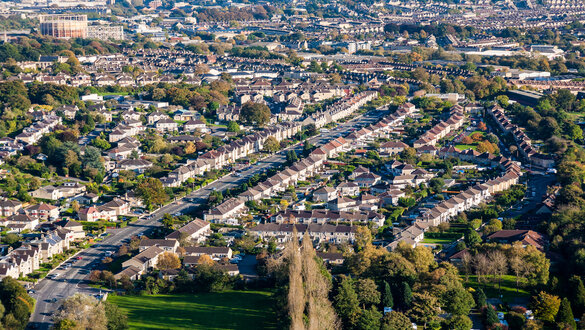Aerial view of a dense suburban neighborhood with rows of houses, tree-lined streets, and a green park in the foreground.