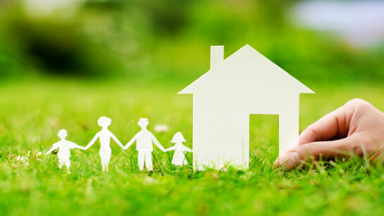 Paper-cut figures of a family holding hands beside a white house on green grass, with a hand nearby placing the house into position.