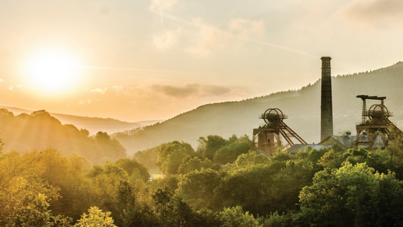 Sunrise over a forested valley with an old industrial complex and tall smokestacks.