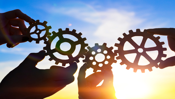Hands joining metal gears against a sunset sky, symbolizing teamwork and collaboration in engineering/production.
