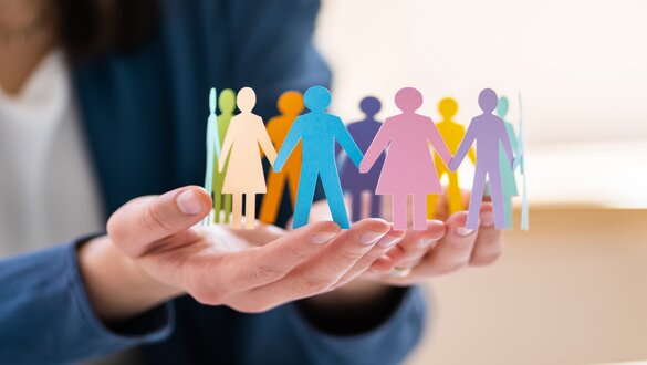 Colorful paper cutout figures of people holding hands, cradled in a pair of hands, symbolizing diversity and unity.