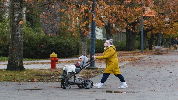 Woman in a yellow coat pushing a baby stroller along a tree-lined park path in autumn leaves.