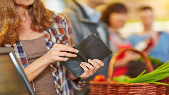Person holding a black wallet while shopping, with a wicker basket of vegetables in the foreground at a market stall