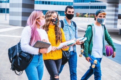 Four students wearing masks walk outside a campus building, carrying notebooks and backpacks, with a basketball visible.