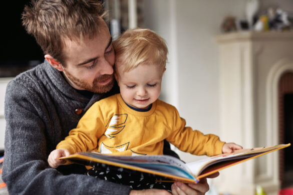 Father reading a colorful picture book with his young son at home, both focused on the pages.