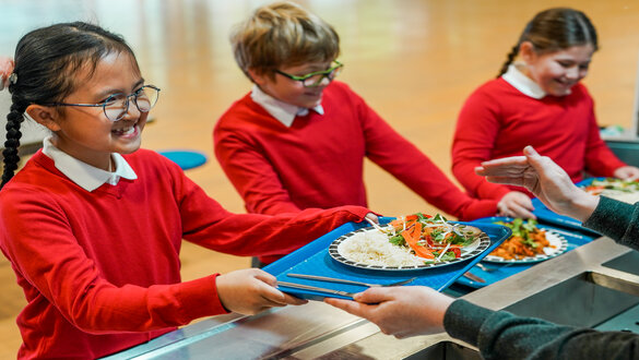 Three smiling students in red uniforms stand at a cafeteria serving counter as they receive trays of food on blue trays.
