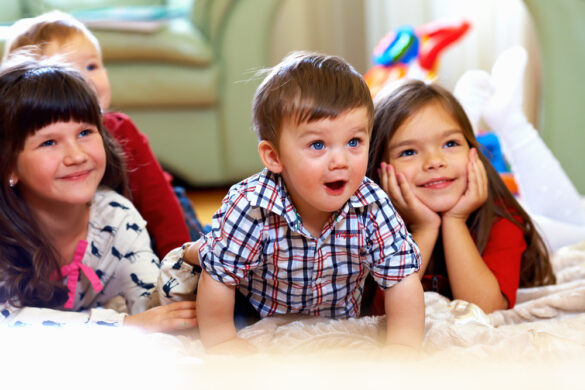 Four young children lying on a soft rug, smiling and watching something off-camera in a cozy indoor setting.