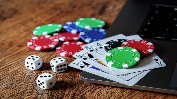 Poker chips, two dice, and playing cards on a wooden table near a laptop.