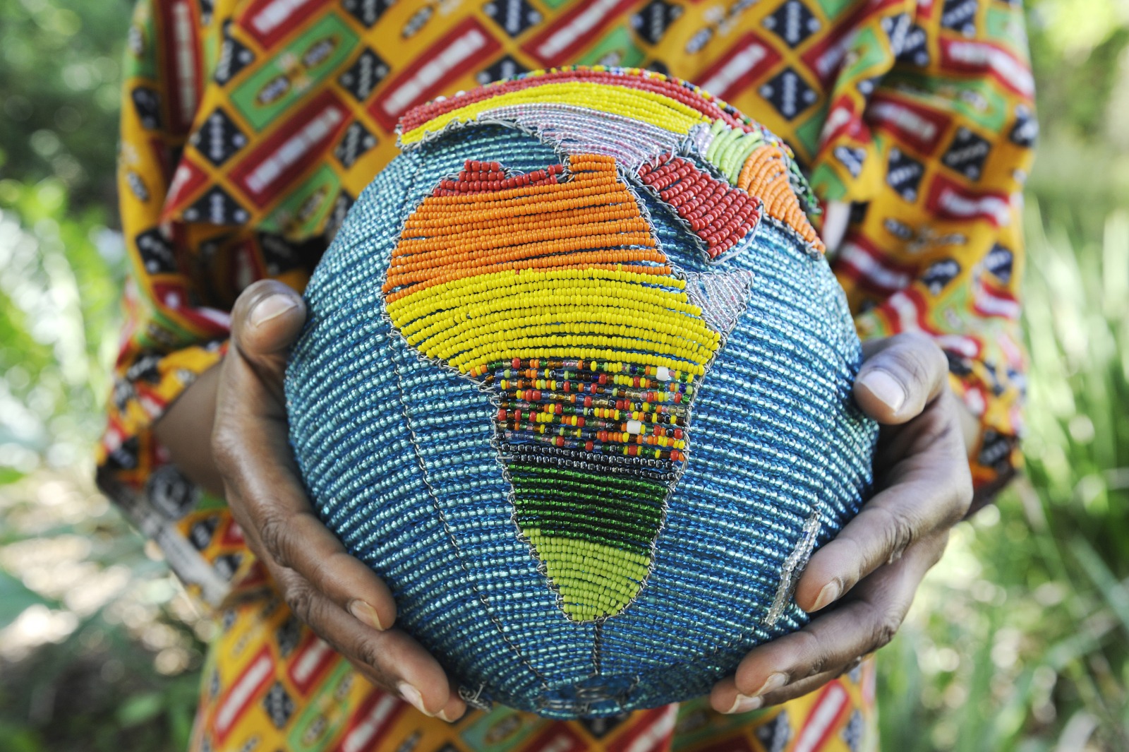 Decorative beaded ball with a multicolored Africa silhouette, held by hands in a patterned garment.